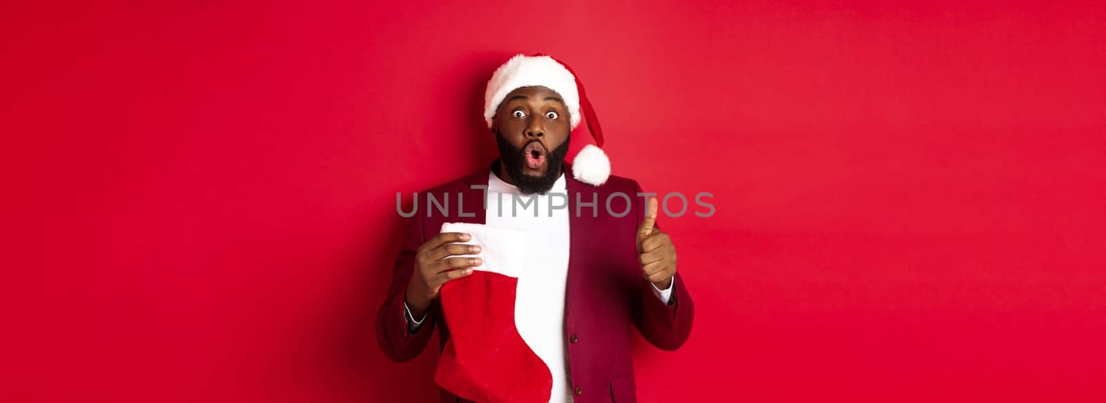 Impressed african american man holding christmas sock with presents, showing thumb up, looking surprised, standing over red background.
