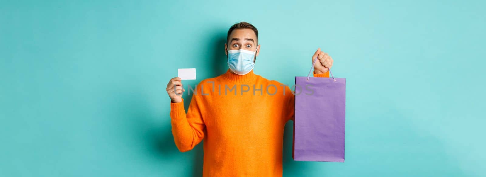 Covid-19, pandemic and lifestyle concept. Happy male customer showing credit card and purple shopping bag, standing over light blue background.