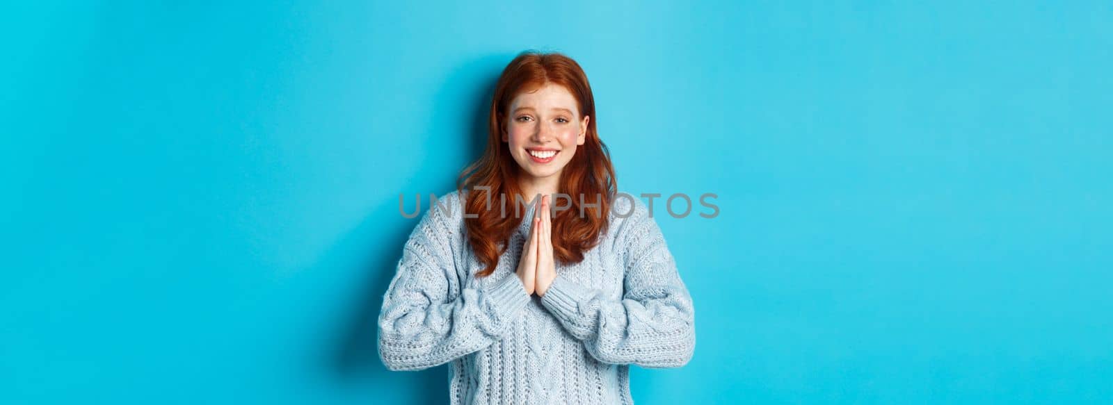 Cute redhead girl saying thank you, smiling and looking at camera, expressing gratitude, standing against blue background.