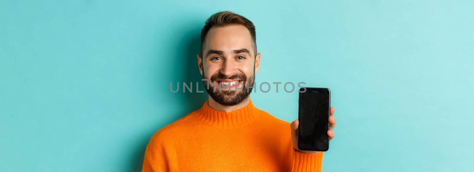 Close-up of handsome bearded guy in orange sweater, showing smartphone screen and smiling, showing promo online, turquoise background.