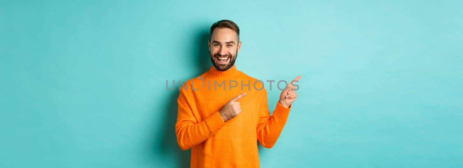 Handsome smiling man pointing fingers left, showing your logo, standing in winter orange sweater, turquoise background.