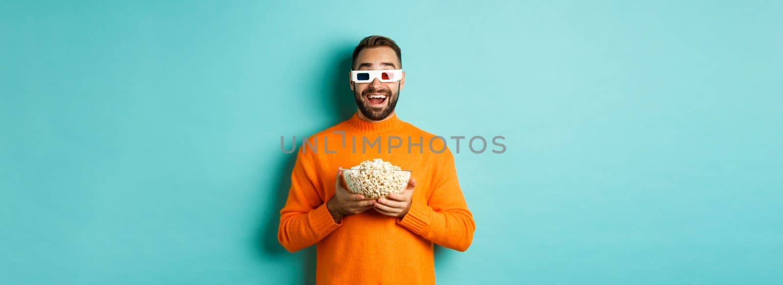 Happy guy watching movies in 3d glasses, eating popcorn and looking at camera, standing over light blue background. Copy space