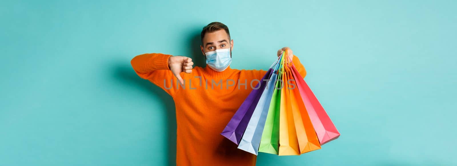 Covid-19, social distancing and lifestyle concept. Disappointed young man in medical mask from coronavirus, showing thumb down and shopping bags.
