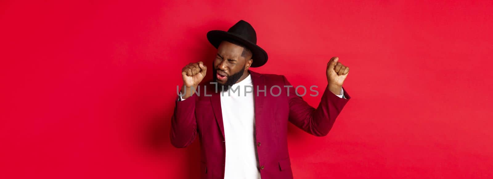 Christmas and people concept. Happy Black man celebrating New Year and dancing, wearing party outfit, red background.