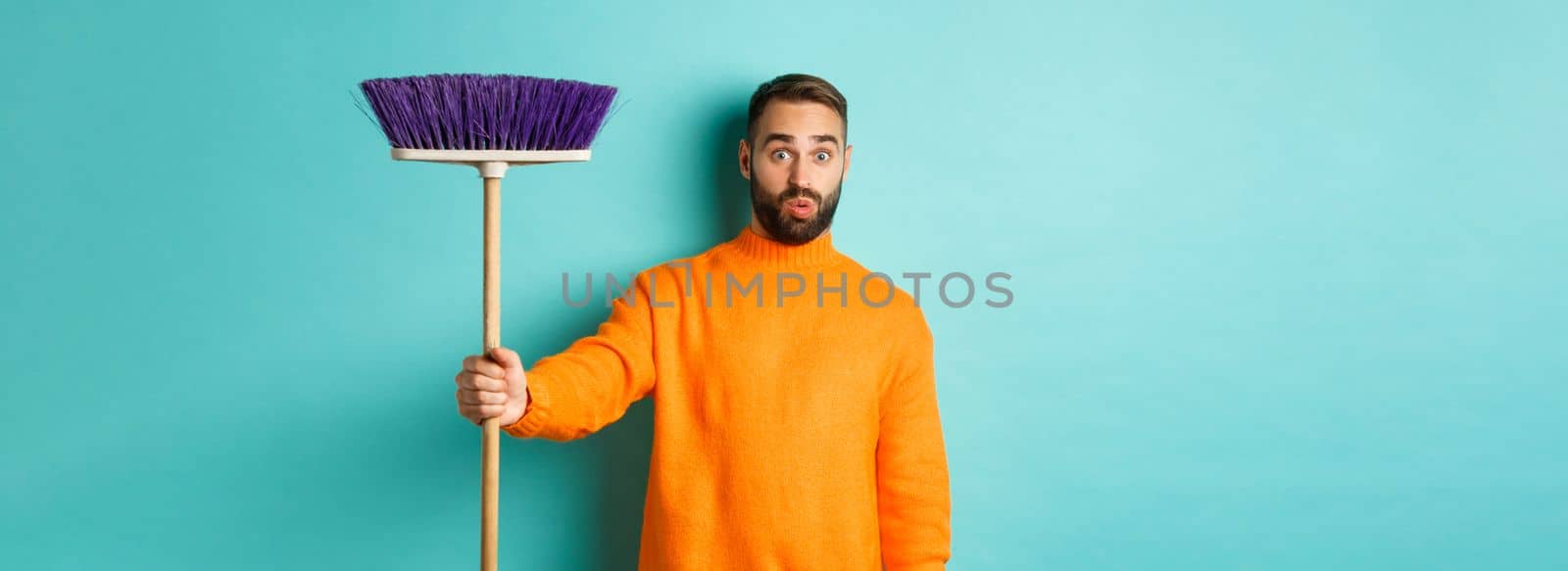 Shocked man receiving broom to do house chores, looking confused, standing over light blue background.