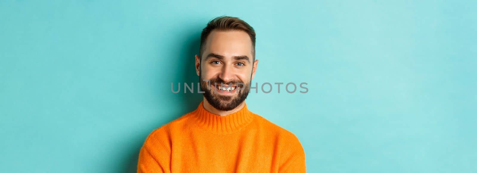 Close-up of handsome caucasian man smiling at camera, looking confident, wearing orange sweater, standing against turquoise background.