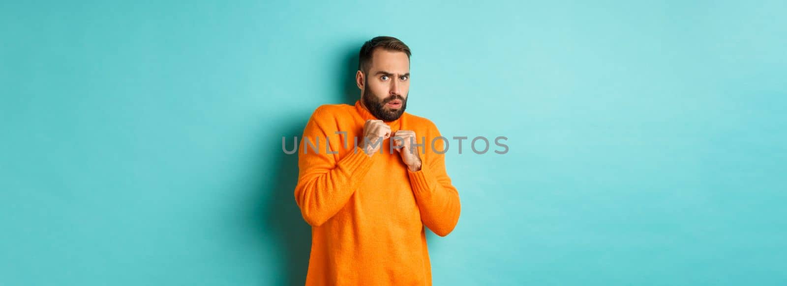 Scared guy jumping startled, looking at something scary, standing in orange sweater, studio background.