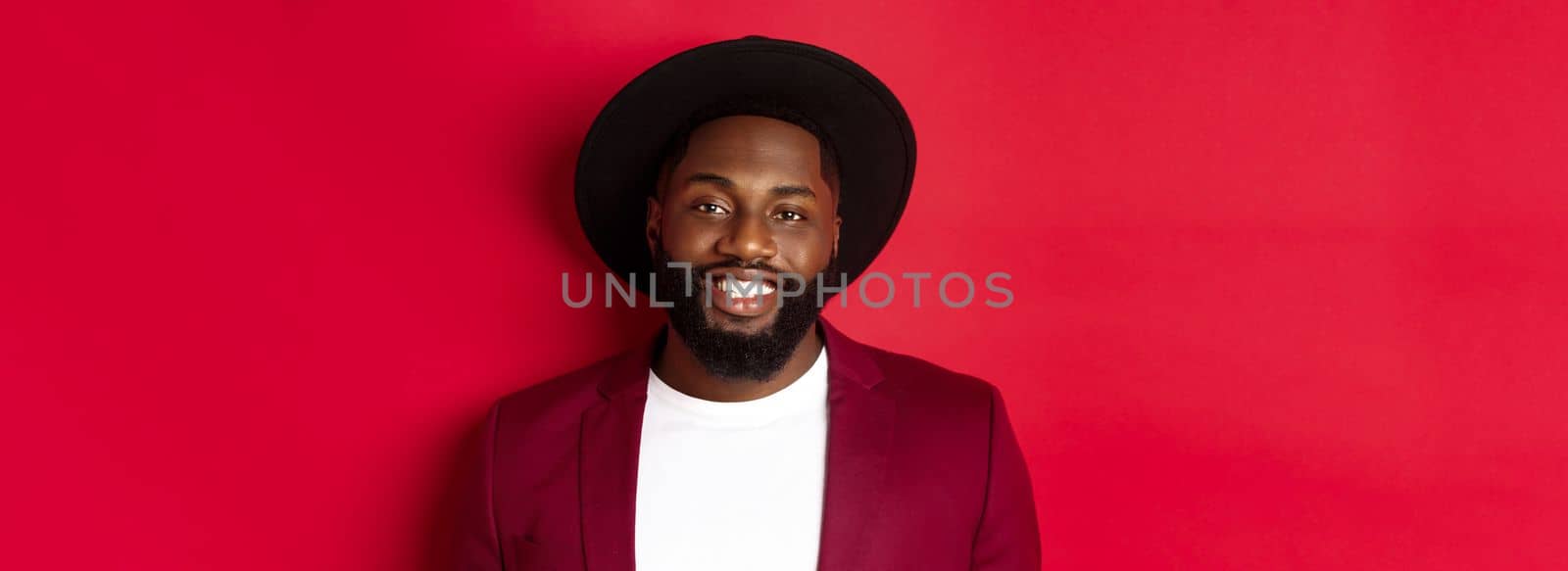 Close-up of handsome african american man with beard, wearing party blazer and stylish hat, smiling at camera, red background.