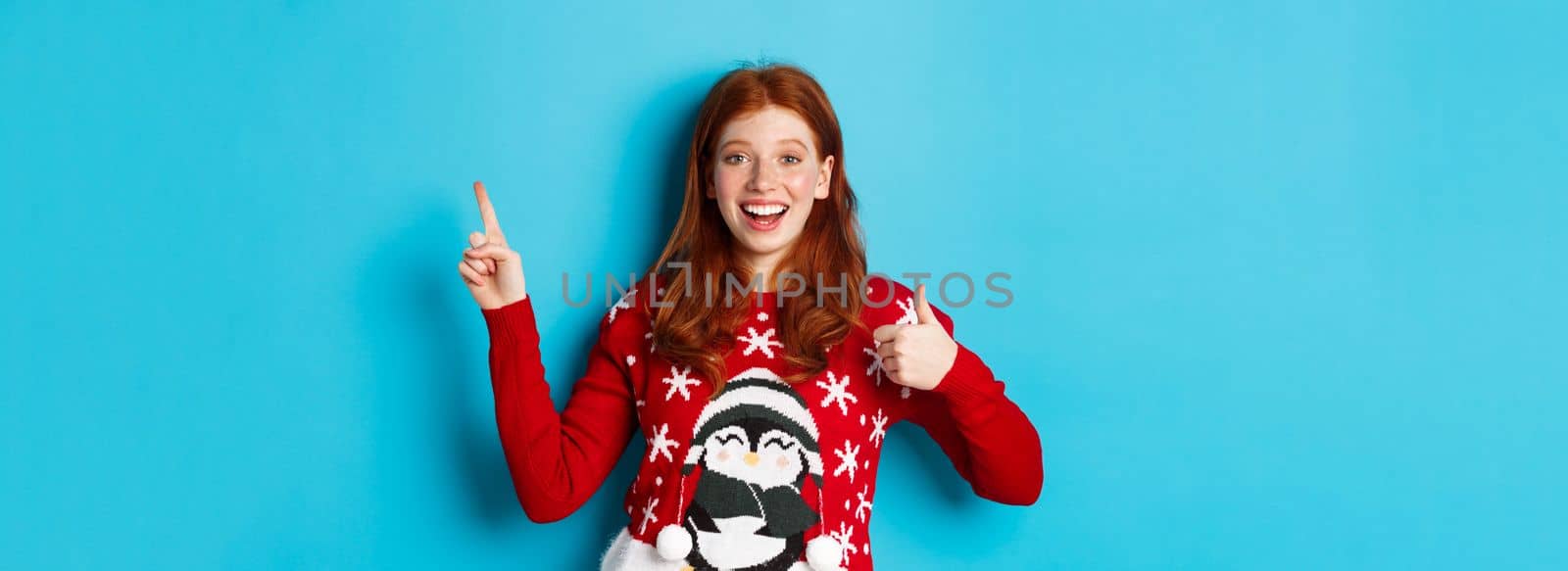 Merry Christmas. Cheerful redhead girl in xmas sweater, pointing finger at upper right corner, showing new year promo and thumbs-up in approval, praise product.