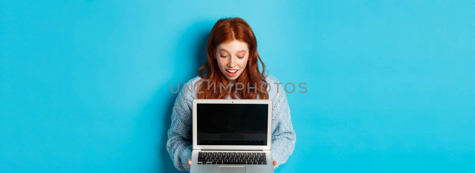 Amazed redhead girl staring at laptop screen and looking impressed, showing computer display, standing over blue background.