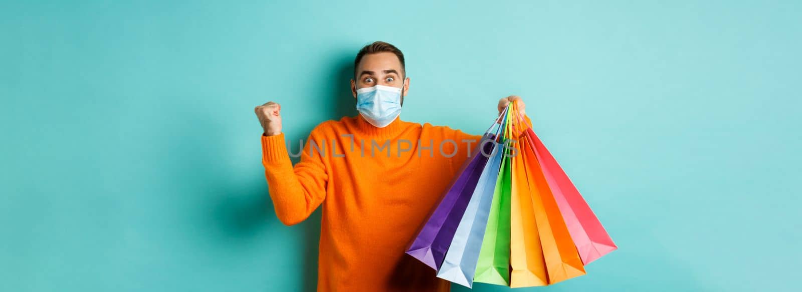 Covid-19, social distancing and lifestyle concept. Excited man in face mask showing shopping bags and rejoicing from discounts, standing over turquoise background.