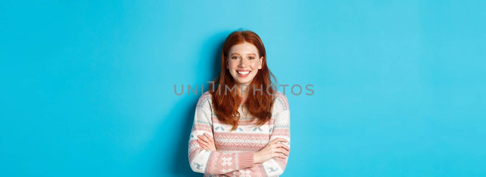 Portrait of happy redhead girl in winter sweater, smiling with arms crossed on chest, standing against blue background.