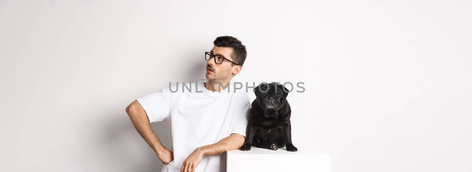 Handsome young man standing near cute black pug, looking right with arrogant expression, standing over white background.