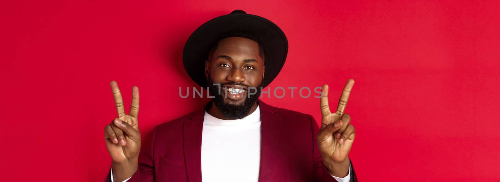 Close-up of handsome bearded Black man showing peace signs and smiling happy, posing for phoro, standing over red background.