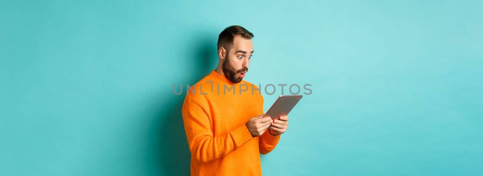 Image of male model in orange sweater staring at digital tablet screen, looking surprised, standing over light blue background.