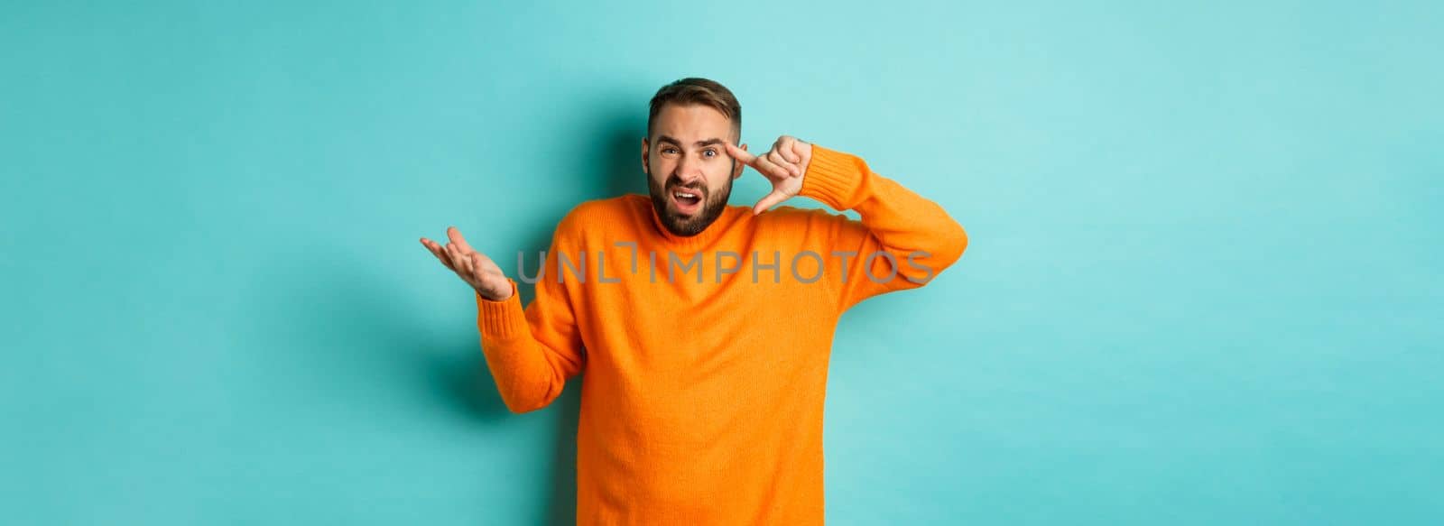 Angry man pointing at head, are you crazy gesture, scolding someone, standing over light blue background. Copy space