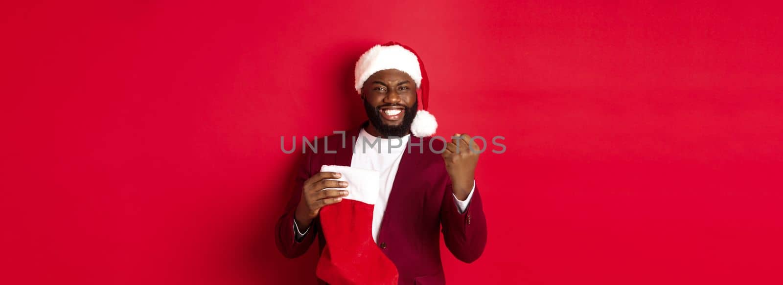 Happy man rejoicing, receive presents in Christmas sock, making fist pump and smiling satisfied, standing over red background.