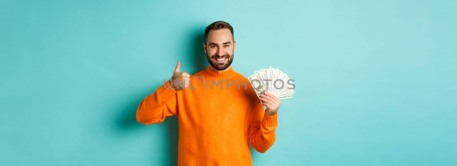Shopping. Happy man showing thumb up and money in dollars, recommending bank loan or credits, standing over light blue background.