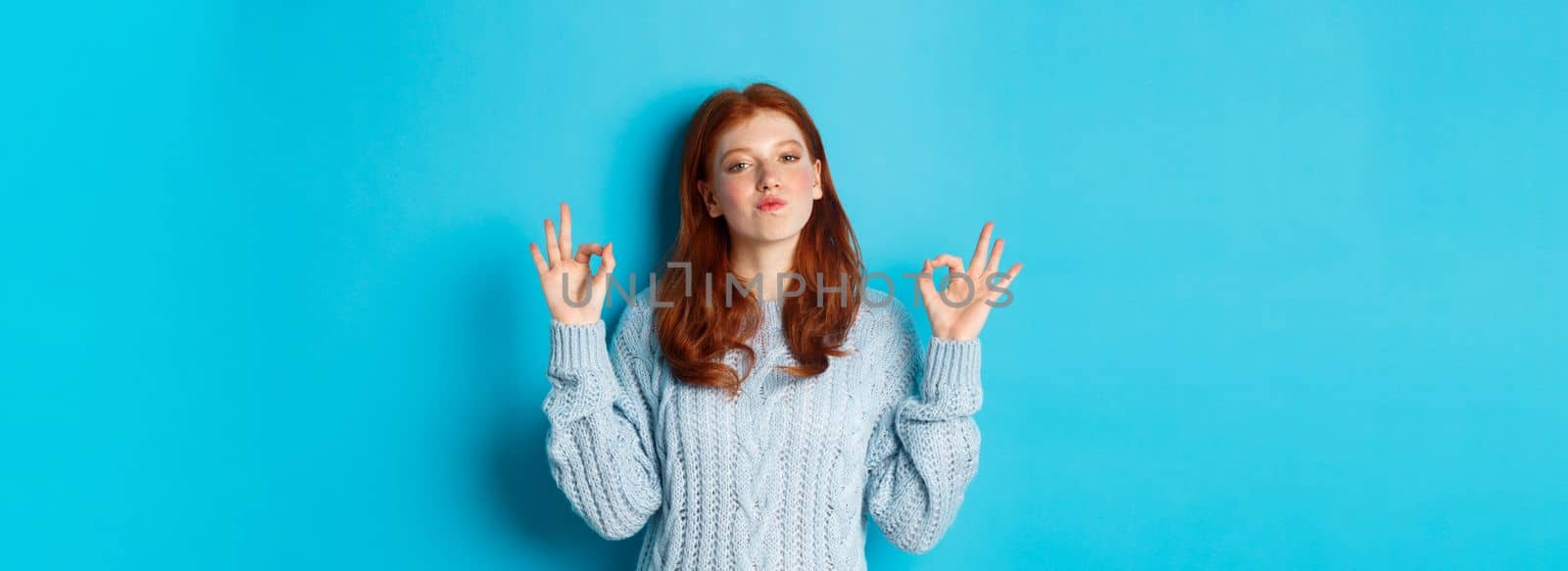 Redhead teenage girl showing okay signs, looking satisfied and proud, agree, give positive answer, praising good choice, standing over blue background.