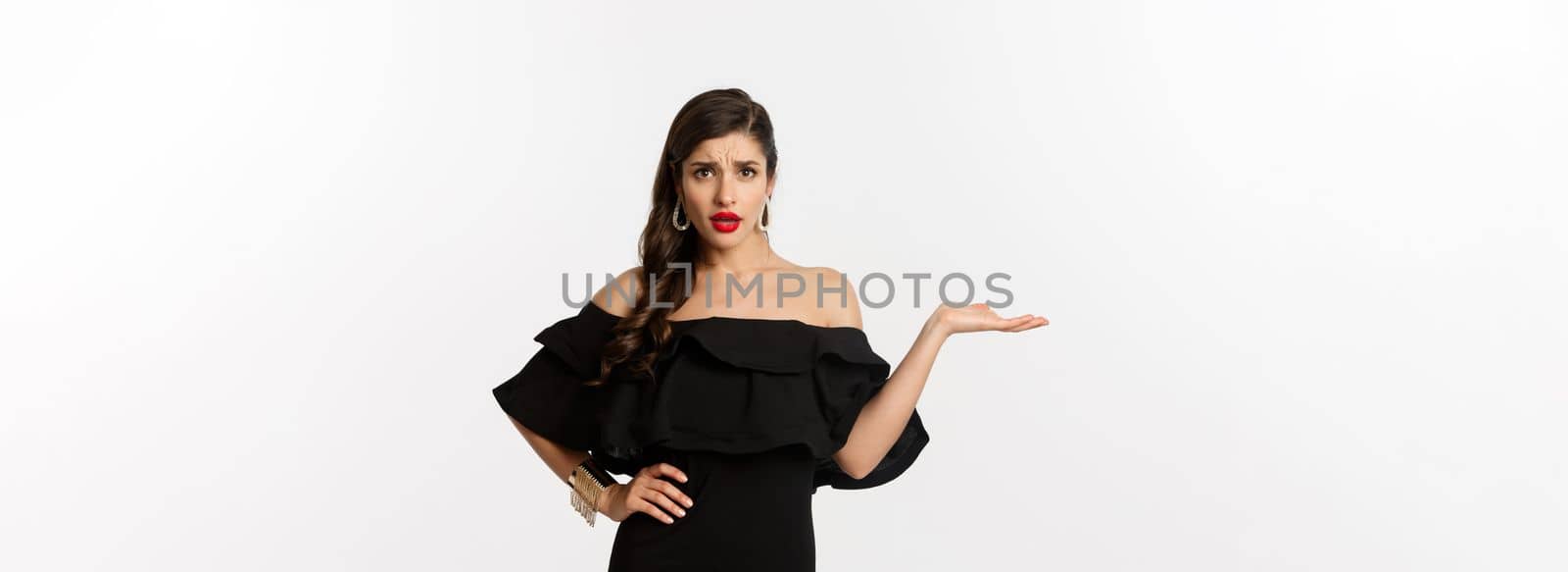 Fashion and beauty. Annoyed woman in black dress raising hand, so what gesture, looking confused at camera, standing bothered over white background.