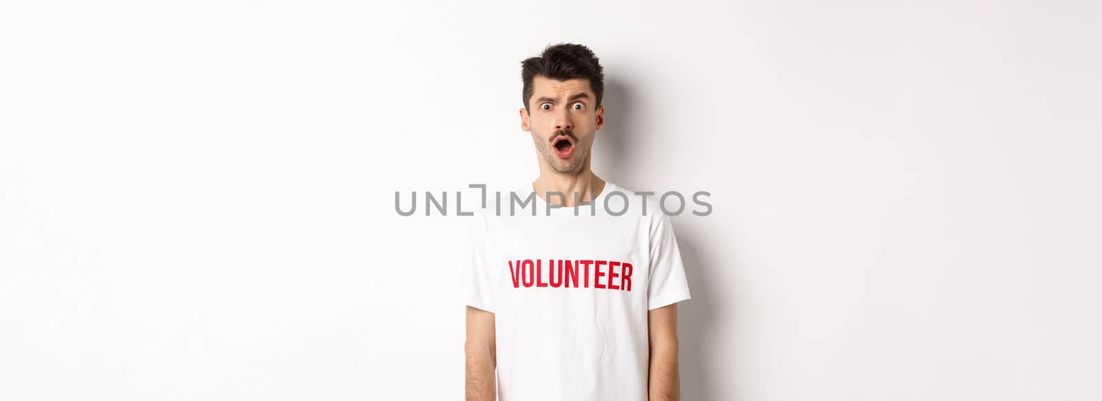 Shocked and confused man in volunteer t-shirt staring at camera speechless, standing against white background.