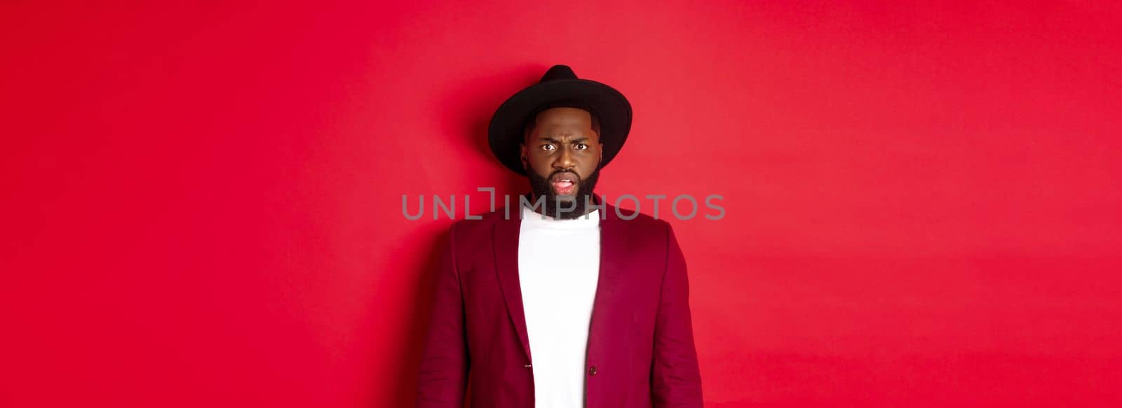 Fashion and party concept. Confused Black man in classy outfit frowning and staring at camera displeased, cant understand, standing over red background.