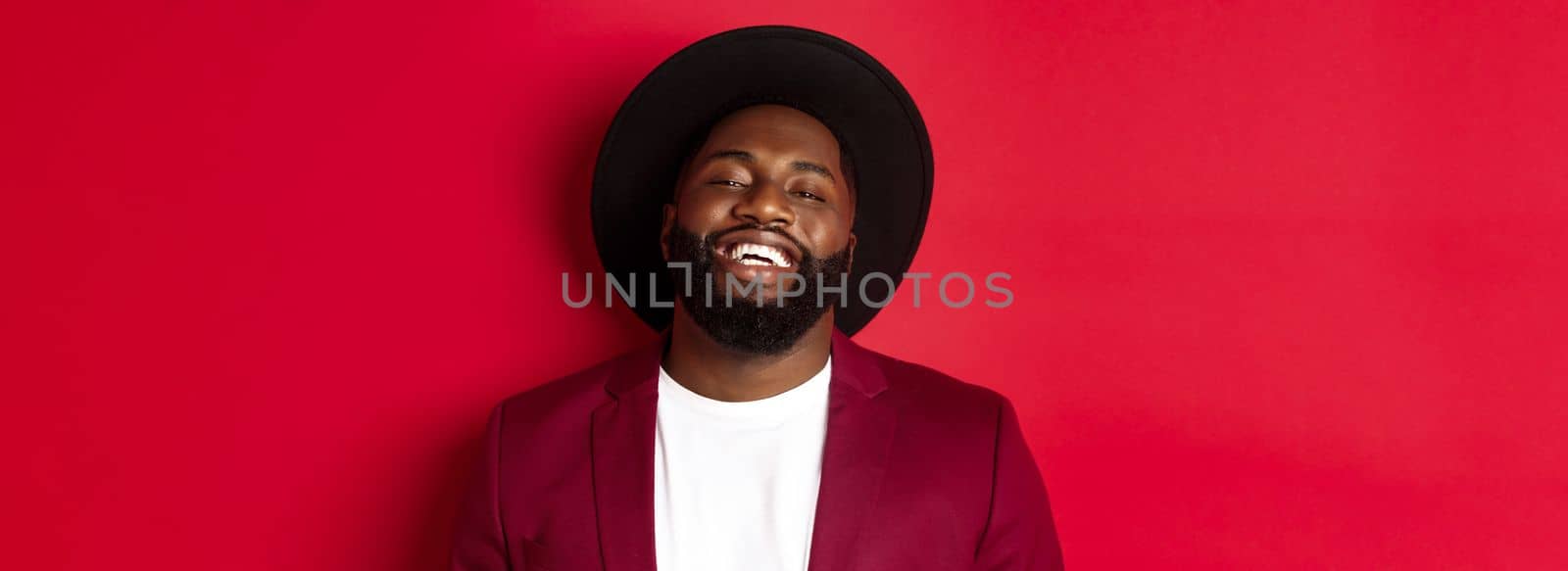 Close-up of happy handsome Black man smiling at you, looking pleased, wearing black hat and blazer, red background.