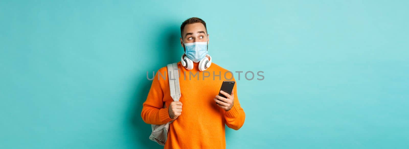 Young man in face mask using mobile phone, holding backpack, staring left amazed, standing against light blue background.