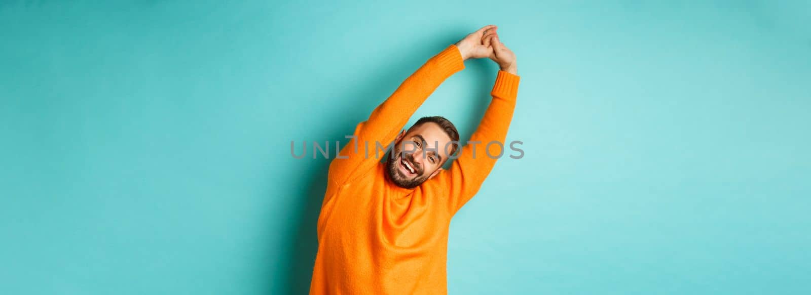 Image of handsome young man stretching hands and smiling after good rest, standing in orange sweater over light blue background.