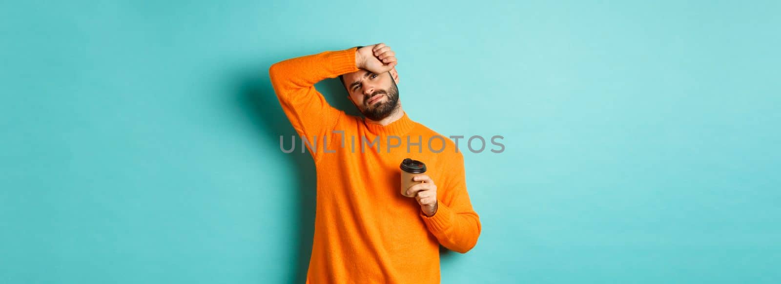 Tired adult man taking break from work and drinking coffee, wipe sweat off forehead and looking exhausted, standing in orange sweater over turquoise background.