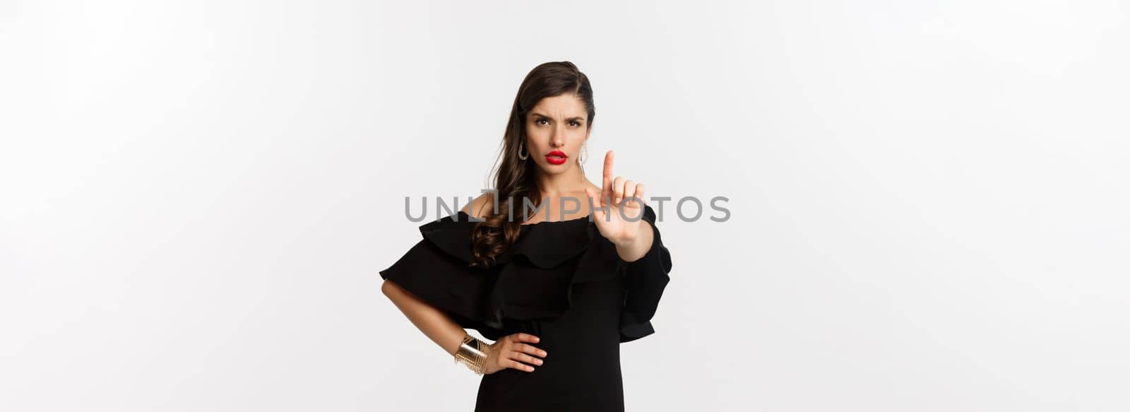 Fashion and beauty. Confident and serious lady in black dress, showing finger in stop gesture, prohibit and disapprove something, standing over white background.