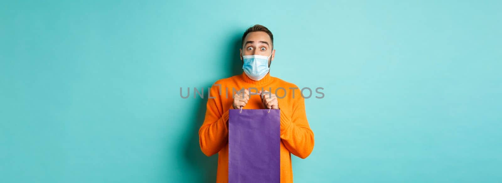 Covid-19, pandemic and lifestyle concept. Surprised man open shopping bag and looking amazed, receiving gift on holiday, standing over turquoise background.