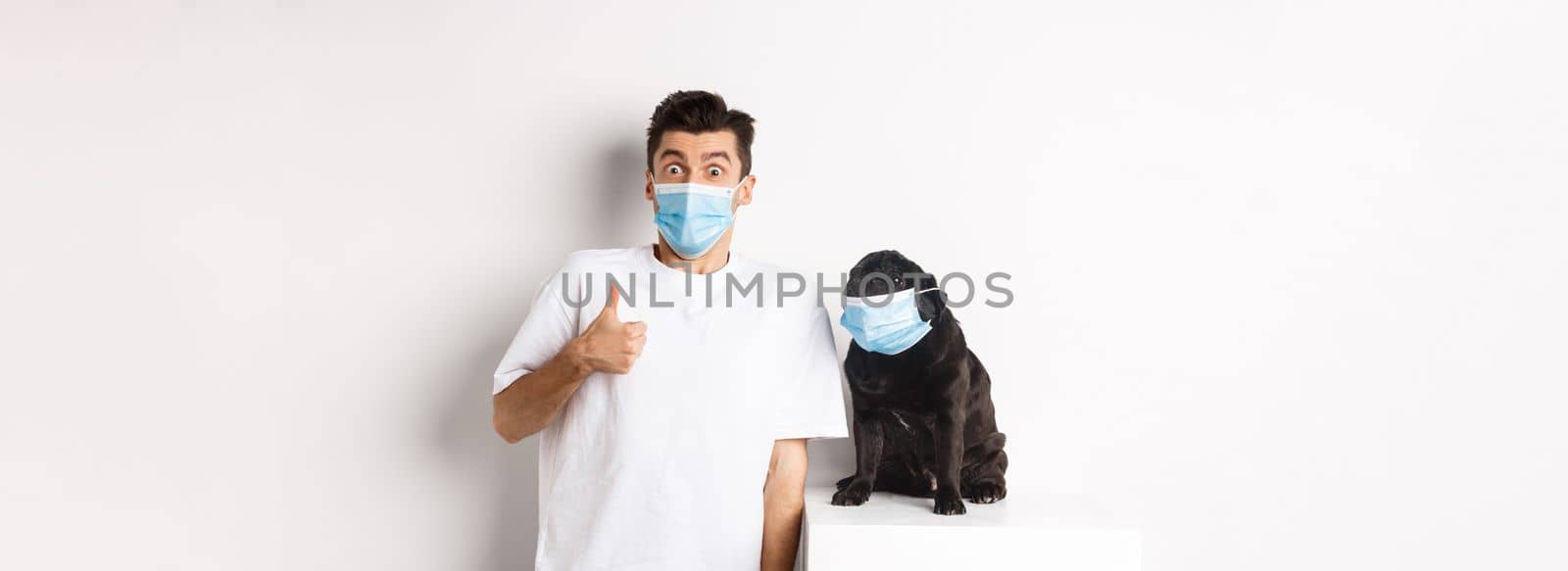 Covid-19, animals and quarantine concept. Image of funny young man and small dog in medical masks, owner showing thumb up in approval or like, white background by Benzoix