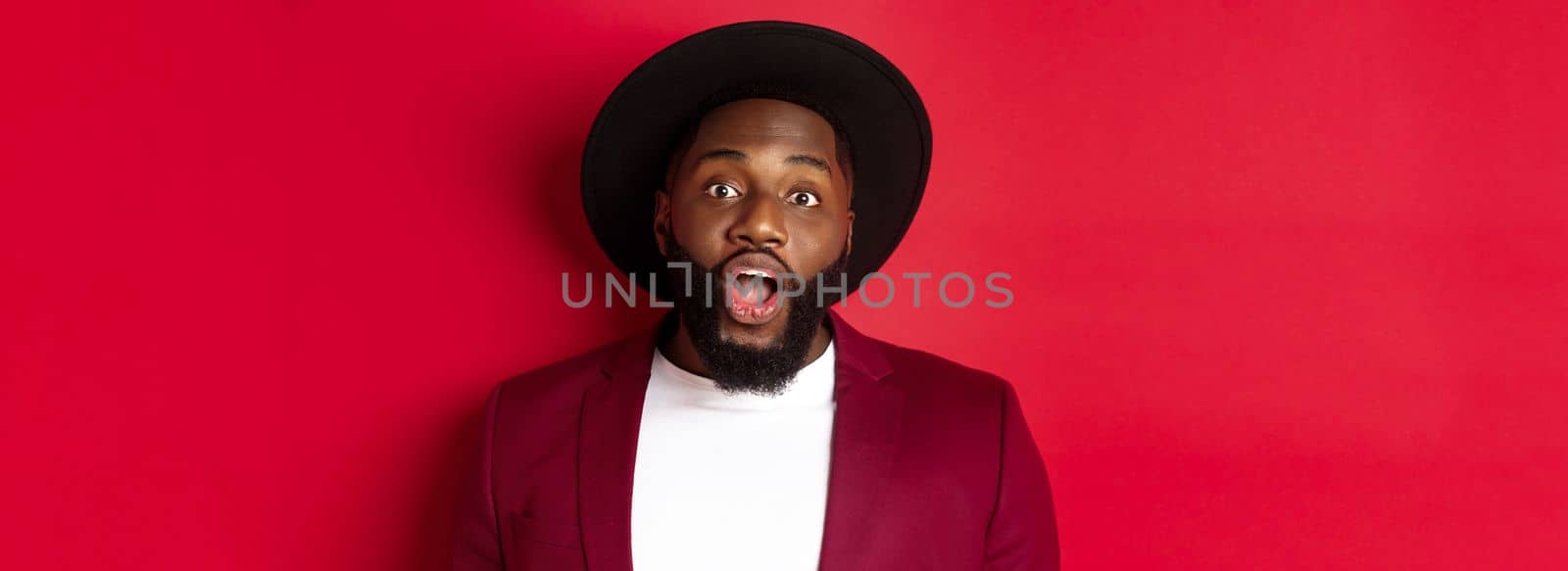Close-up of impressed Black man with beard staring at camera, drop jaw and looking amazed, standing over red background.