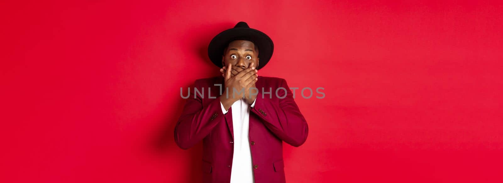 Christmas shopping and people concept. Shocked black man looking startled at camera, covering mouth with hands, standing against red background.