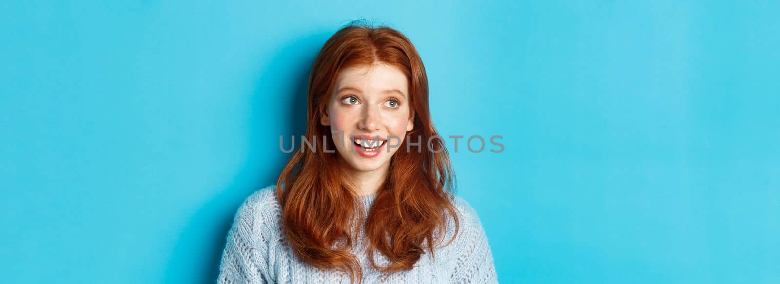 Close-up of beautiful redhead girl in sweater, looking at upper left corner promo logo and smiling, standing over blue background by Benzoix