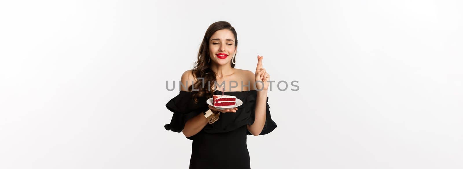 Celebration and party concept. Hopeful and dreamy woman making wish on birthday cake, cross fingers and smiling happy, standing over white background.