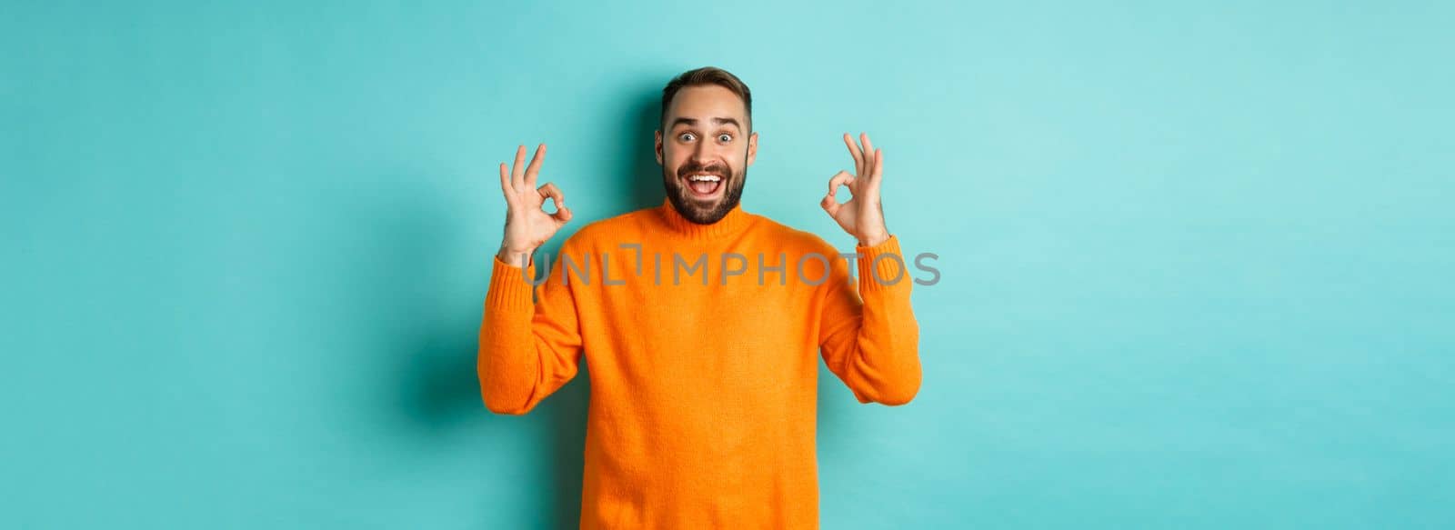 Impressed man praising something awesome, showing okay signs and looking amazed, standing in orange sweater at light blue background.