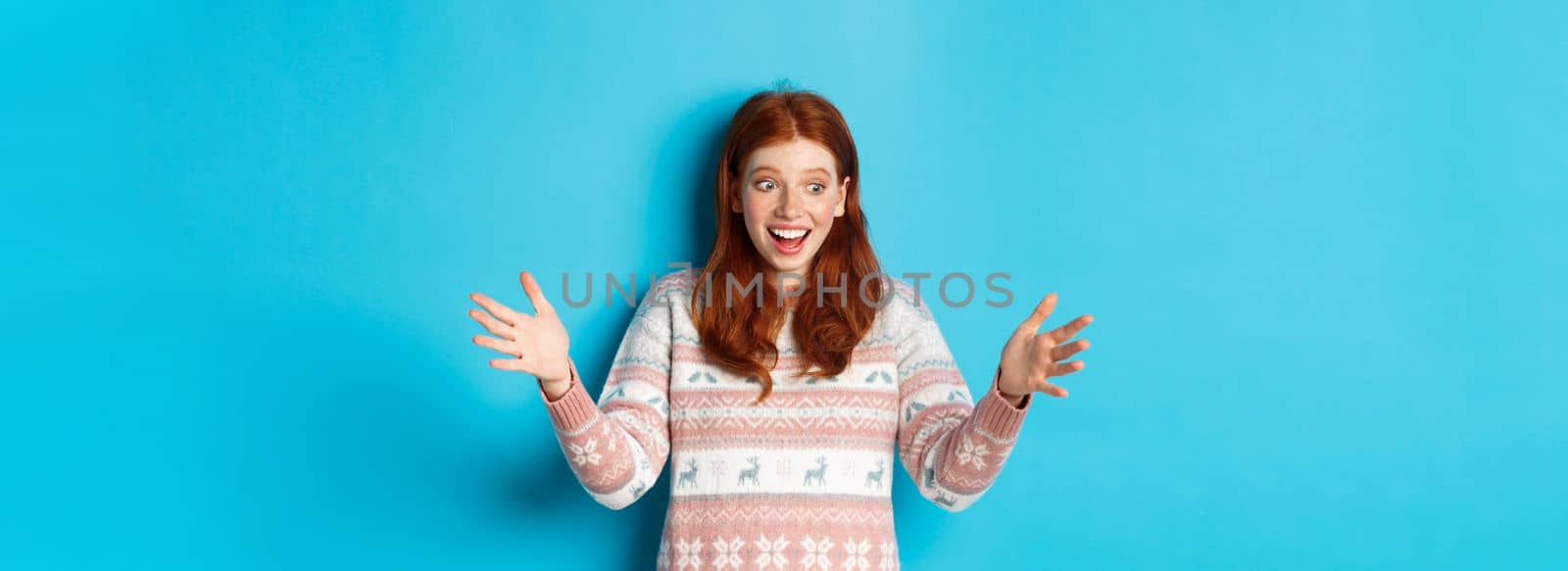 Image of happy redhead girl holding big size object, looking amazed and smiling, receiving Christmas gift, standing over blue background.