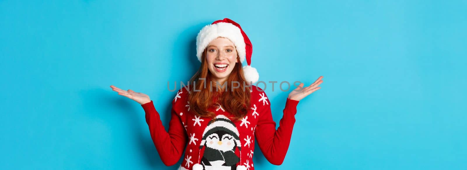 Happy holidays and Christmas concept. Cheerful redhead girl in Santa hat and xmas sweater, raising hands on copy spaces, holding something on blue background.