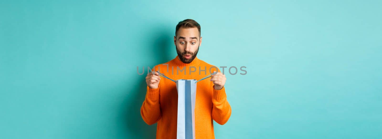 Surprised man open shopping bag and looking amazed, receiving gift on holiday, standing over turquoise background.