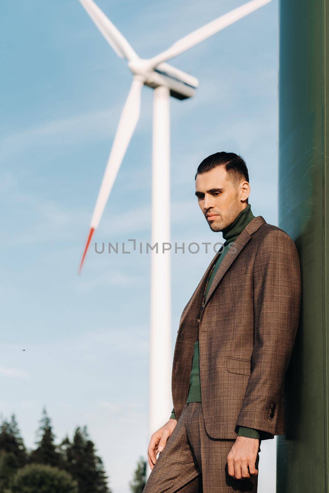 A man in a business suit with a green Golf shirt stands next to a windmill against the background of the field and the blue sky.Businessman near the windmills.Modern concept of the future. by Lobachad