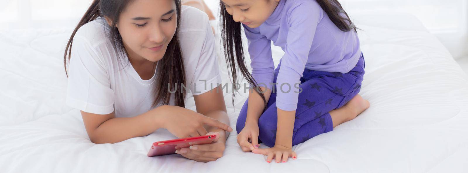Young asian mother and little daughter looking tablet computer together for learning and education on bed in bedroom, mom and girl watching tablet to internet at home, family and technology concept.