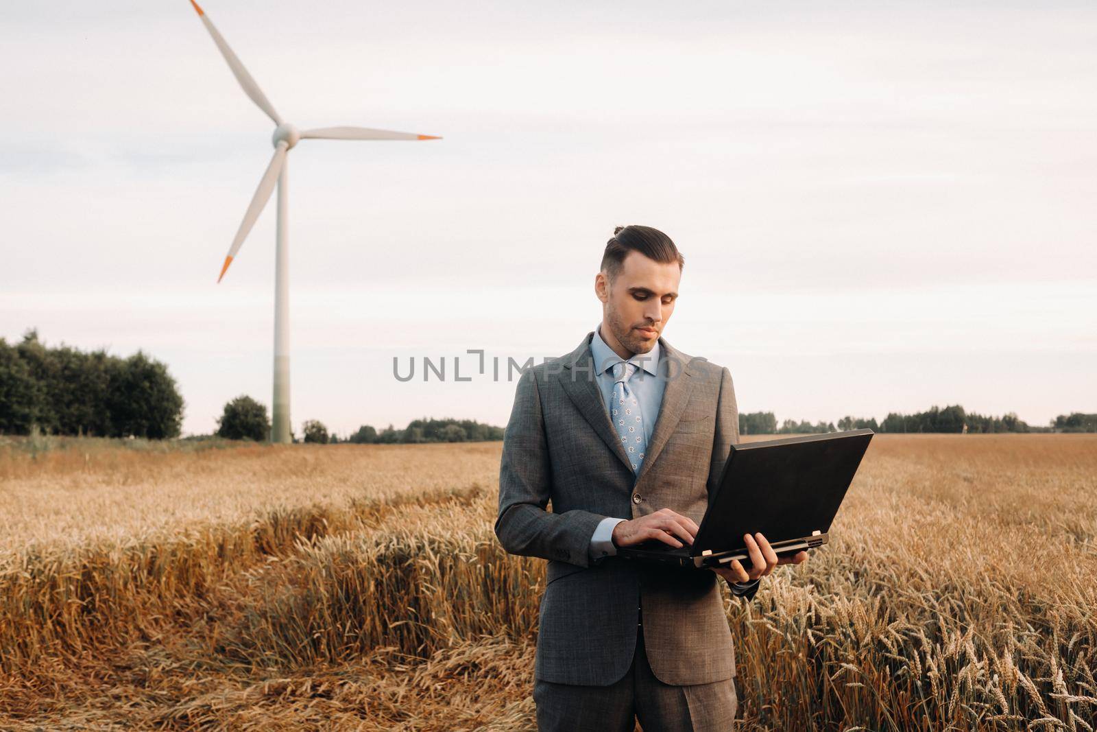 Portrait of a businessman in a suit holding a laptop in a field of wheat against the background of a windmill and the evening sky.