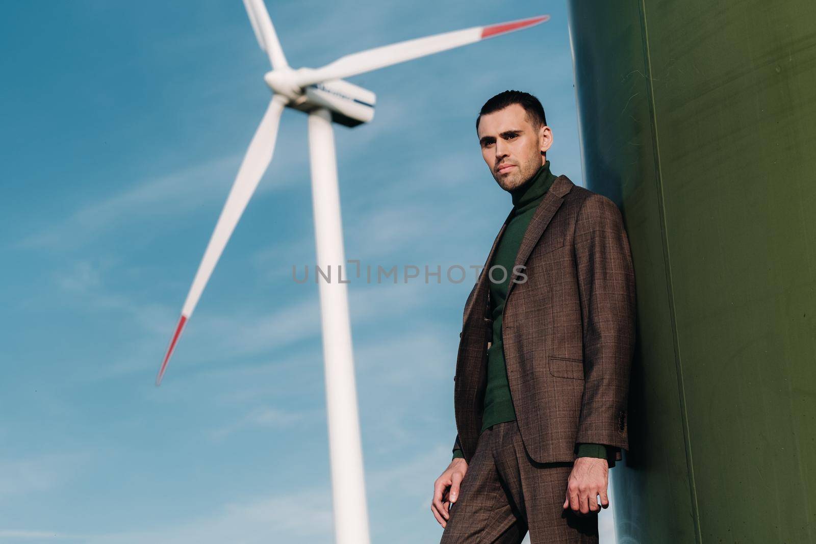 A man in a business suit with a green Golf shirt stands next to a windmill against the background of the field and the blue sky.Businessman near the windmills.Modern concept of the future