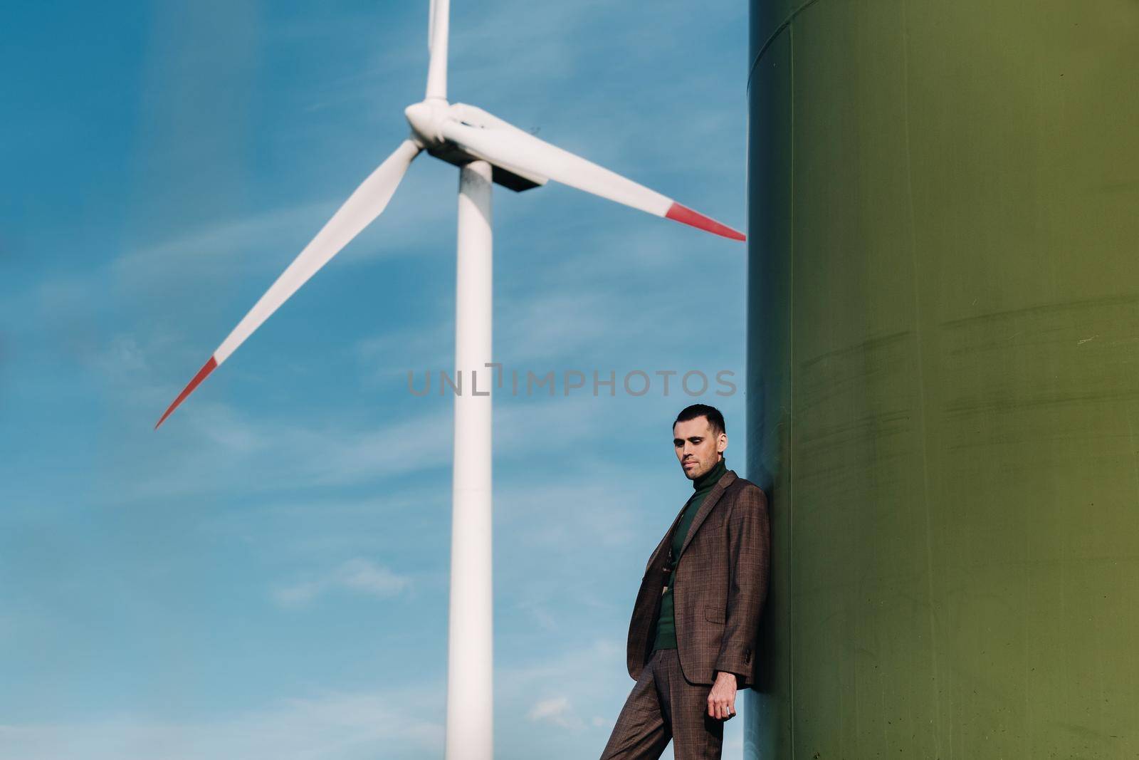 A man in a business suit with a green Golf shirt stands next to a windmill against the background of the field and the blue sky.Businessman near the windmills.Modern concept of the future. by Lobachad