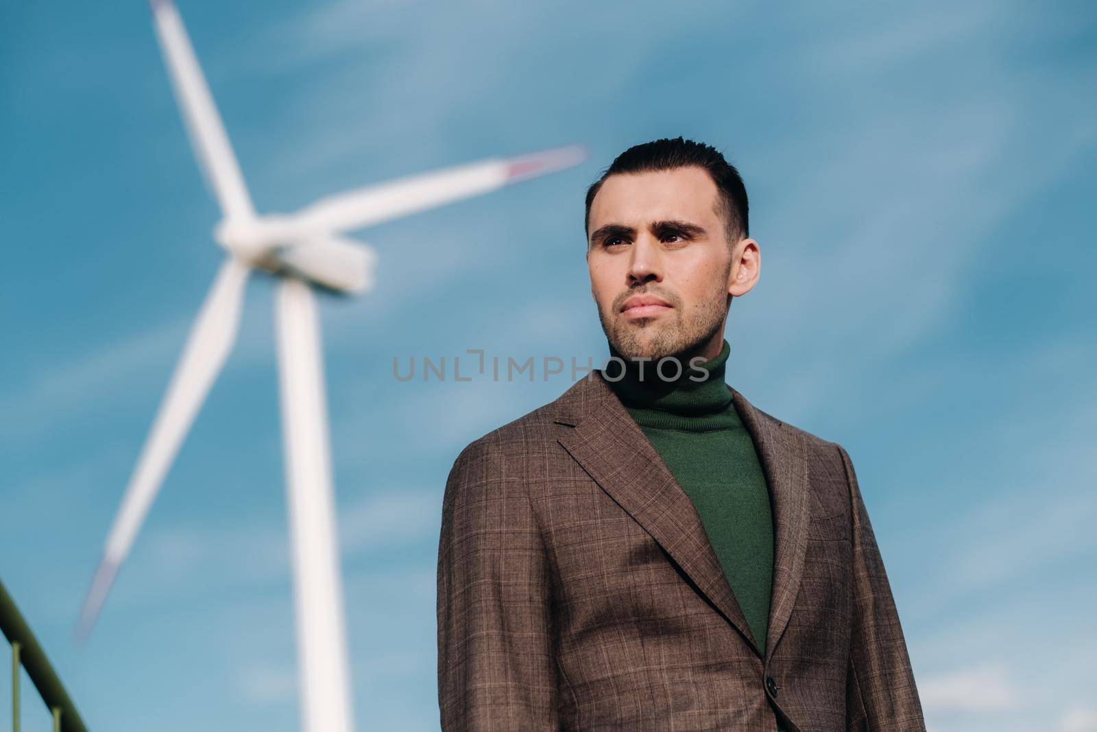 A man in a business suit with a green Golf shirt stands next to a windmill against the background of the field and the blue sky.Businessman near the windmills.Modern concept of the future