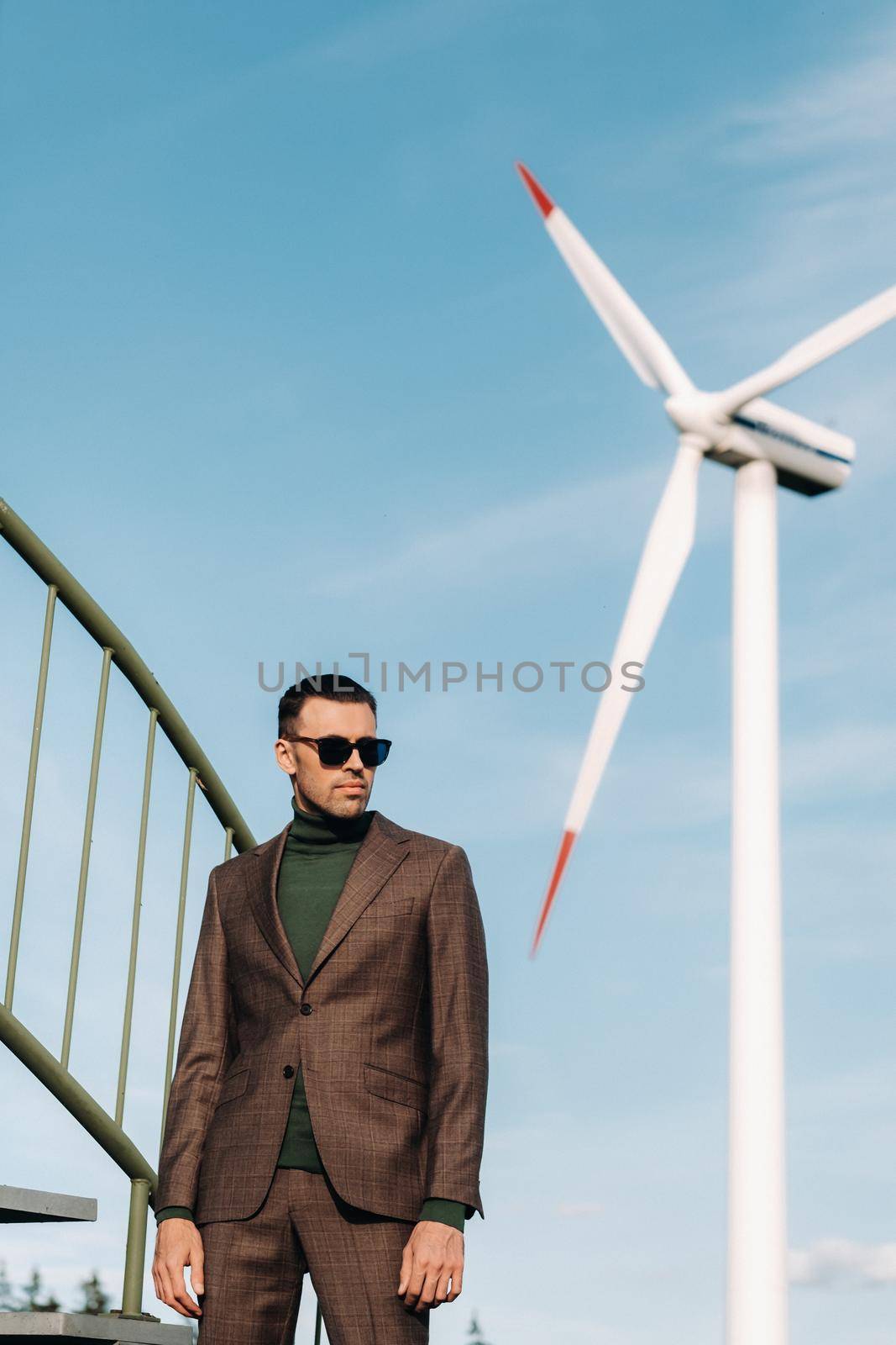 A man in a business suit with a green Golf shirt stands next to a windmill against the background of the field and the blue sky.Businessman near the windmills.Modern concept of the future. by Lobachad