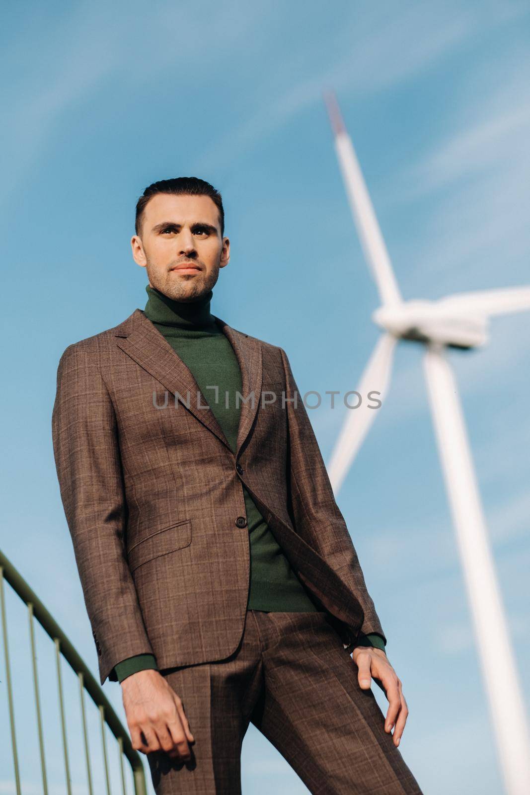 A man in a business suit with a green Golf shirt stands next to a windmill against the background of the field and the blue sky.Businessman near the windmills.Modern concept of the future