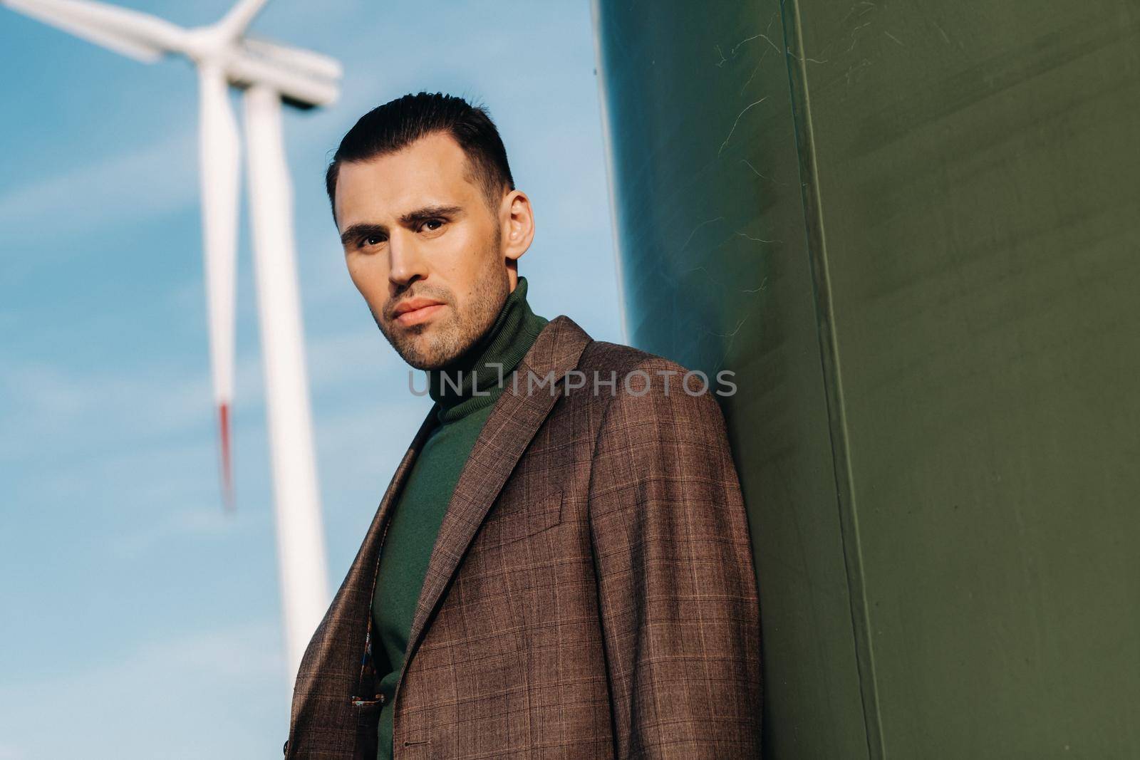 A man in a business suit with a green Golf shirt stands next to a windmill against the background of the field and the blue sky.Businessman near the windmills.Modern concept of the future. by Lobachad